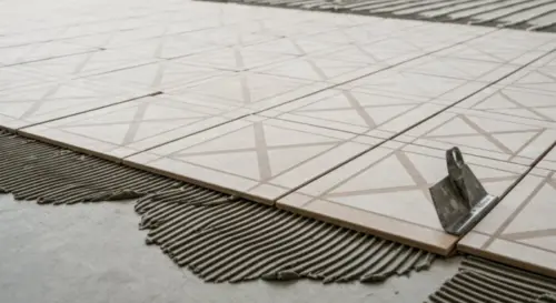 A detailed close-up photo of porcelain floor tiles being installed over underfloor heating pipes, showing flexible adhesive application with a notched trowel on a concrete subfloor, with thermal expan