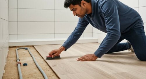 A close-up, photorealistic image of a professional installer laying vinyl plank flooring in a modern bathroom with underfloor heating pipes visible in the subfloor, showing proper expansion gaps along