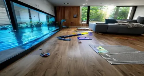 A realistic photo of a modern living room with a large aquarium on one side, showing vinyl flooring installation in progress near the tank. A radiant heating system diagram is visible on a tablet near