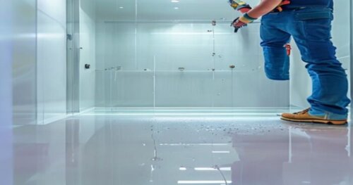 Photorealistic close-up of a glass shower enclosure with memory effect hinges being installed in a modern bathroom. The floor is glossy epoxy resin, and the walls are white drywall. A worker is using