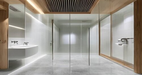 A modern bathroom with a frameless glass shower screen installed over a seamless liquid porcelain floor. The floor has a matte finish, light grey color. The walls are white tiles. In the ceiling, a ve