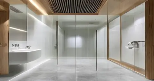 A modern bathroom with a frameless glass shower screen installed over a seamless liquid porcelain floor. The floor has a matte finish, light grey color. The walls are white tiles. In the ceiling, a ve