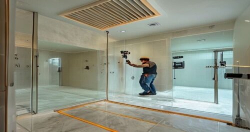 A realistic photo of a modern bathroom with a glass shower enclosure being installed on a porcelain tile floor. The floor has visible underfloor heating pipes marked with tape. A worker is using a dri
