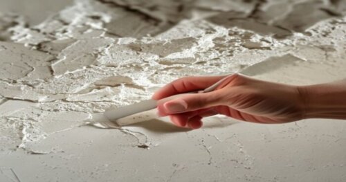 A close-up photorealistic image of a hand applying white joint compound over a mesh tape on a crack in a plaster ceiling. The background shows a textured ceiling surface. The hand is using a putty kni