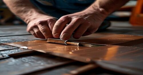 Close-up of a craftsman's hands cutting a square section of floating laminate floor with a utility knife, revealing a copper pipe with a small leak underneath. The floor has underfloor heating pipes v
