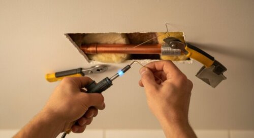 A close-up, photorealistic image of a professional plumber's hands working on a copper pipe repair inside a small hole cut in a white plaster false ceiling. The hands are holding a soldering torch and
