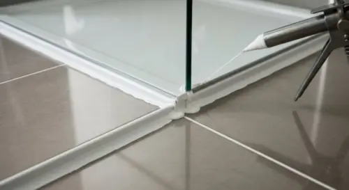 A close-up photo of a bathroom shower screen installed on a porcelain tile floor with underfloor heating. The joint between the glass screen and the tile is being sealed with a white silicone sealant