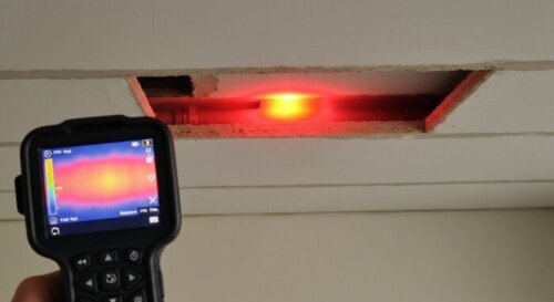 A realistic photo of a thermal camera showing a hot spot on a ceiling, indicating a water leak in a PPR pipe hidden behind a plasterboard false ceiling. The camera display is visible, with a clear col