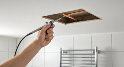 A close-up photorealistic image of a plumber's hands repairing a copper pipe inside a small access hole in a plasterboard ceiling. The hands are holding a propane torch and soldering the pipe joint. T