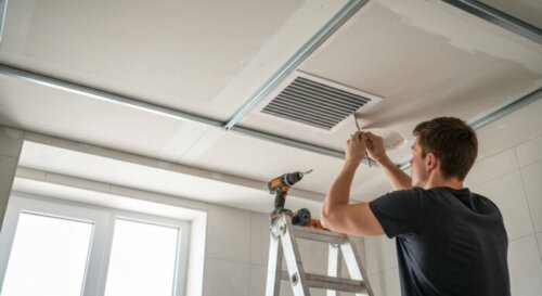 Photorealistic close-up of a bathroom ceiling with a suspended plasterboard false ceiling being installed, showing metal profiles, plasterboard panels, and a ventilation duct. A worker is cutting a ho