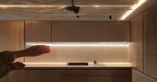 Close-up photo of hands installing a sleek LED strip under a modern kitchen cabinet, with a plasterboard ceiling and hidden cables, realistic lighting, no text.