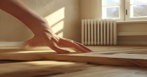 Close-up view of a hand applying beige elastic wood filler into a gap between two floating floorboards, with a heat radiator on the wall in the background to suggest underfloor heating, natural daylig