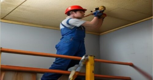A worker in blue overalls installing red PEX pipes on a ceiling with yellow insulation foam boards, using clips and a rail system, before covering with plasterboard. The ceiling has a laser level line